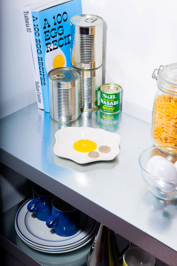 A kitchen shelf holds stacked tins, a wasabi tin, a blue book A 100 Egg Recipes, a pasta jar, two blue cups, a bowl of eggs, and the DOIY Fast Food Fried Egg Trinket Tray by DOIY for charming retro style.