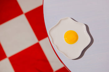 The DOIY Fast Food Fried Egg Trinket Tray by DOIY, featuring a white tray with a yellow center shaped like a fried egg, adds a playful vintage touch to your space.