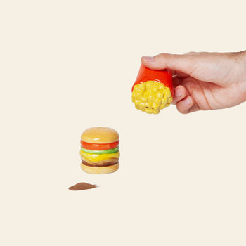 A hand holds the DOIY Fast Food Fries & Burger Salt & Pepper Set—by DOIY—featuring a French fries-shaped salt shaker and a burger-shaped pepper shaker, all styled like vintage decor on a simple, light background.