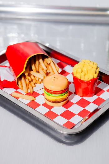 A metal tray holds fries in a red container, the DOIY Fast Food Fries & Burger Salt & Pepper Set, and a red cup with yellow topping—all on red-and-white checkered paper, perfect for vintage decor fans.