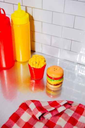 Two condiment bottles, one red and one yellow, sit on a metal counter by a white tiled wall. In front is the DOIY Fast Food Fries & Burger Salt & Pepper Set from DOIY, with retro style and a red-and-white checkered cloth nearby.
