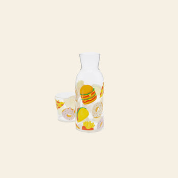 The Stacked Store DOIY Fast Food Jar & Glass Set features a retro carafe and tumbler with playful food illustrations—burger, lemon, hot dog, pizza, taco, croissant—evoking a fun diner vibe against a simple light background.