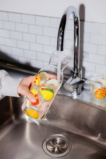 A person fills the Stacked Store DOIY Fast Food Jar from the Jar & Glass Set with water at a kitchen sink, as a matching glass from the set rests on the stainless steel basin.