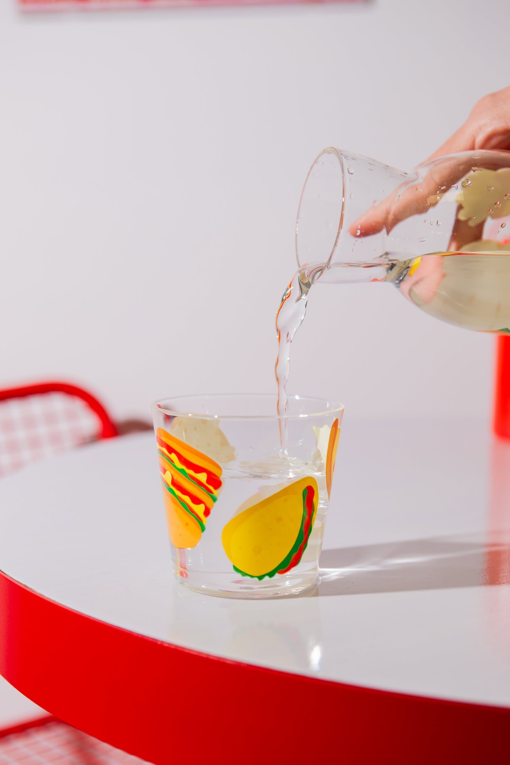 A hand pours water from the Stacked Store DOIY Fast Food Jar & Glass Set into a taco-illustrated glass, placed on a white table with red edges and a red chair in the background.