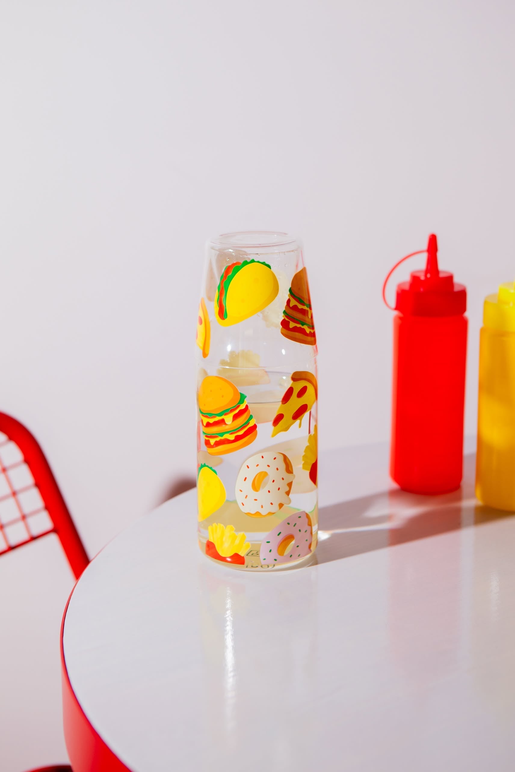 The DOIY Fast Food Jar & Glass Set by Stacked Store, featuring colorful burger, taco, pizza, and donut designs, is displayed on a white table with red and yellow squeeze bottles; a red wire chair appears in the background.