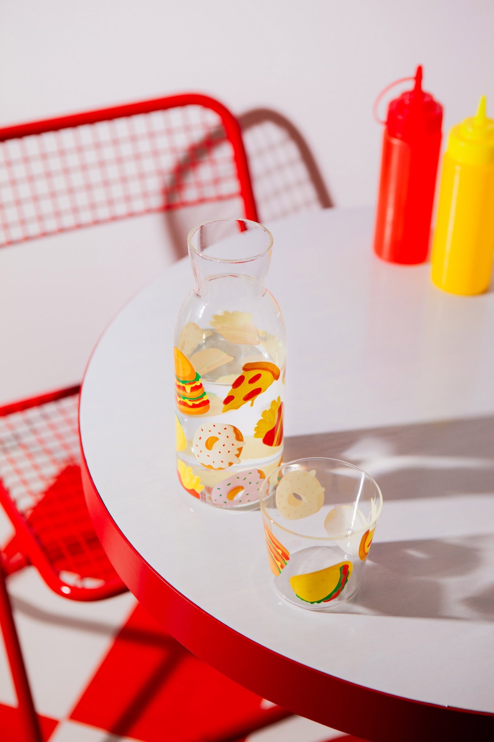 The DOIY Fast Food Jar & Glass Set from Stacked Store, featuring colorful food illustrations, sits on a red-edged table beside red and yellow condiment bottles and a red wire chair, evoking classic diner style.