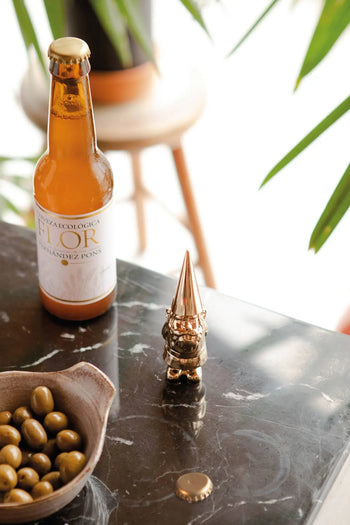 A bottle of beer, the DOIY Gnome Bottle Opener by DOIY, a bowl of green olives, and a bottle cap sit on a dark marble table with green plants and a stool in the background.