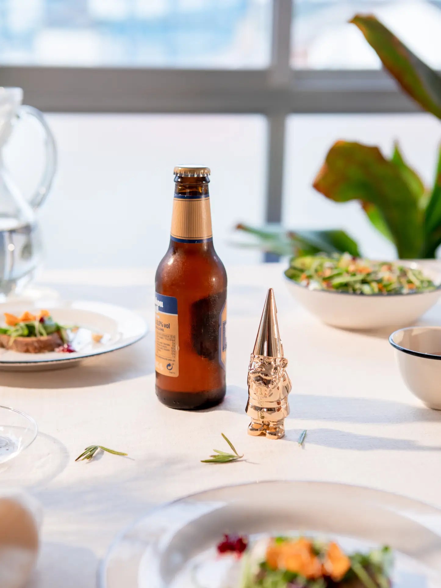 A DOIY Gnome Bottle Opener by DOIY and a brown beer bottle sit on a white tablecloth with plates of food and salad, while a plant and bright window create a lively background.