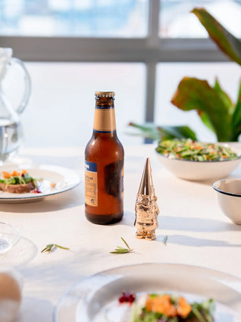 A DOIY Gnome Bottle Opener by DOIY and a brown beer bottle sit on a white tablecloth with plates of food and salad, while a plant and bright window create a lively background.