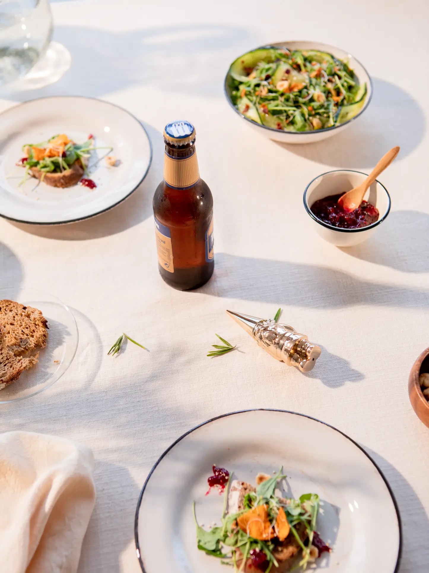 A sunlit table features salad-topped bread, cranberry sauce, a bottle of beer opened with the DOIY Gnome Bottle Opener by DOIY, a salad bowl, sliced bread, and a decorative corkscrew on a white tablecloth.