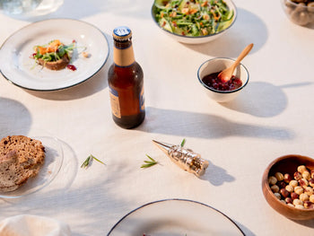 A table set for a meal features a beer bottle, salad, bread, mixed nuts, jam with a wooden spoon, and the DOIY Gnome Bottle Opener by DOIY as the charming centerpiece on a crisp white tablecloth.