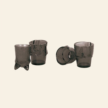 The DOIY Kitty Glasses set includes four smoky gray glasses with unique, crumpled shapes. Arranged in pairs on a beige background, two stand upright and two lie on their sides, creating eye-catching decorative pieces.