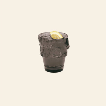 A stackable, dark DOIY Kitty Glass from the DOIY Kitty Glasses Set of 4, filled and garnished with a lemon wedge, stands out against a plain light beige background.