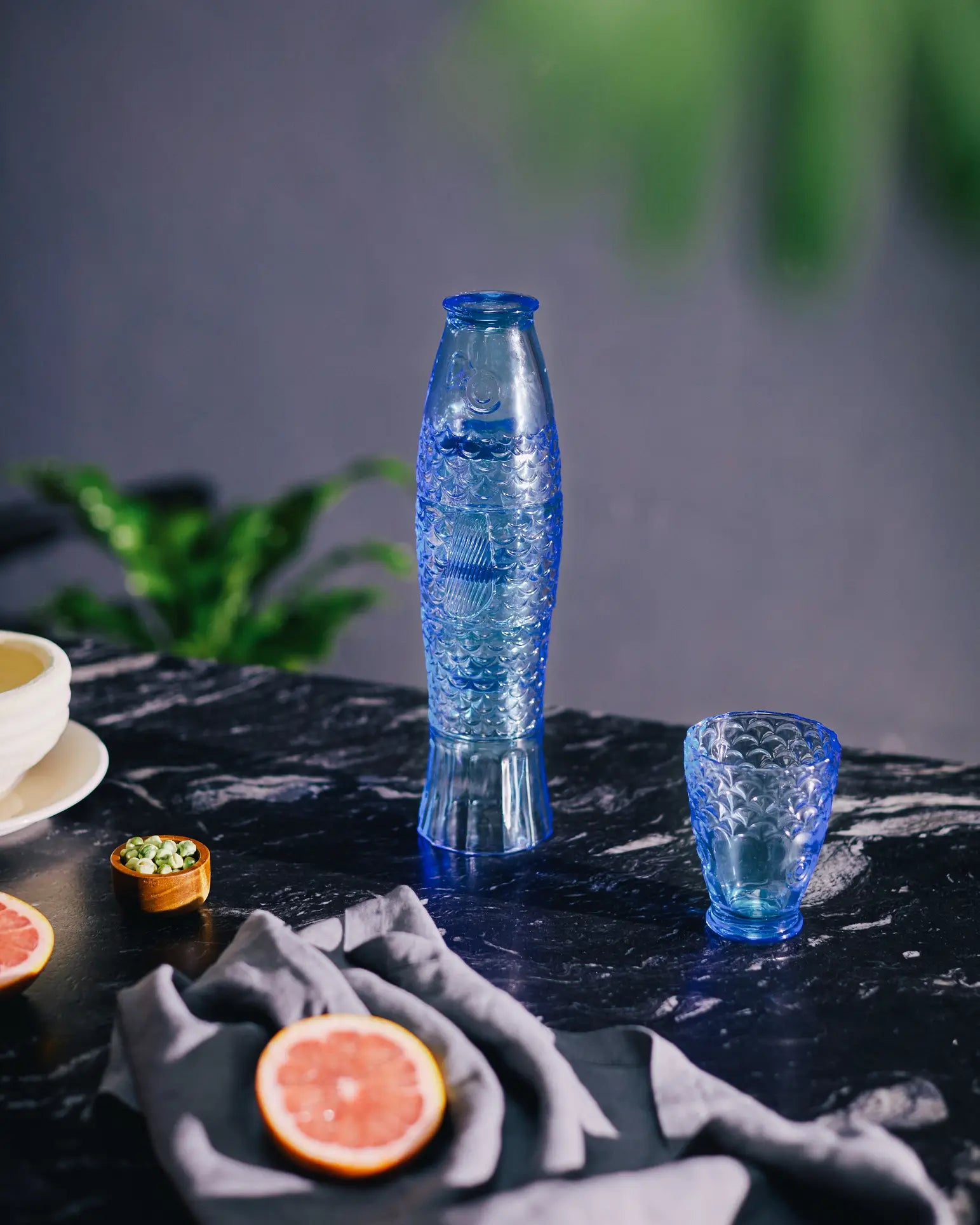 The DOIY Koifish Glasses, Set of 4 by DOIY are displayed on a dark marble surface, paired with a matching glass and surrounded by grapefruit slices, a small bowl, and greenery in the background.