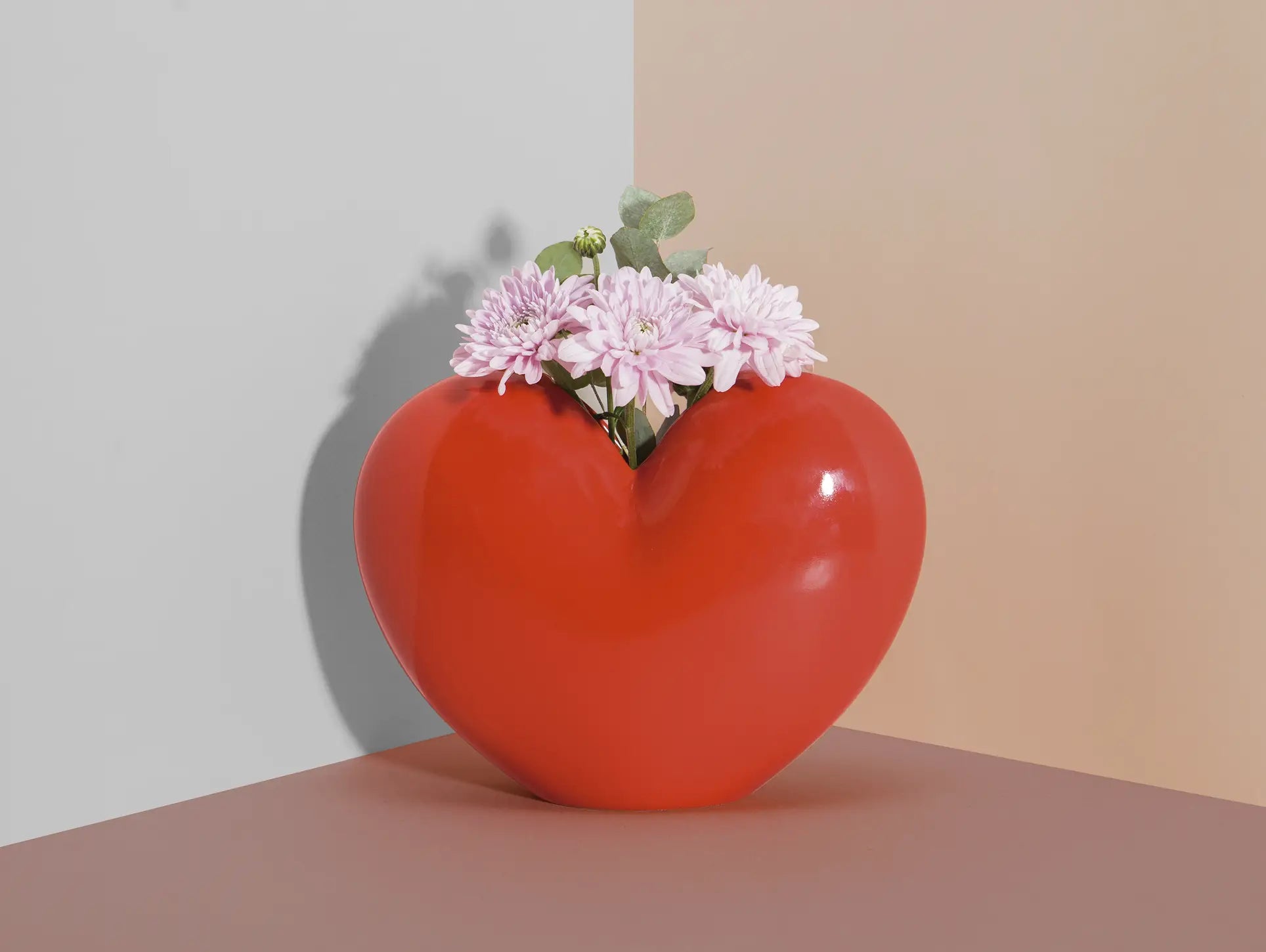 The DOIY Love Vase by DOIY features a shiny red heart-shaped design holding pink flowers with green leaves, beautifully displayed where gray and beige walls meet—an elegant and sentimental décor piece.