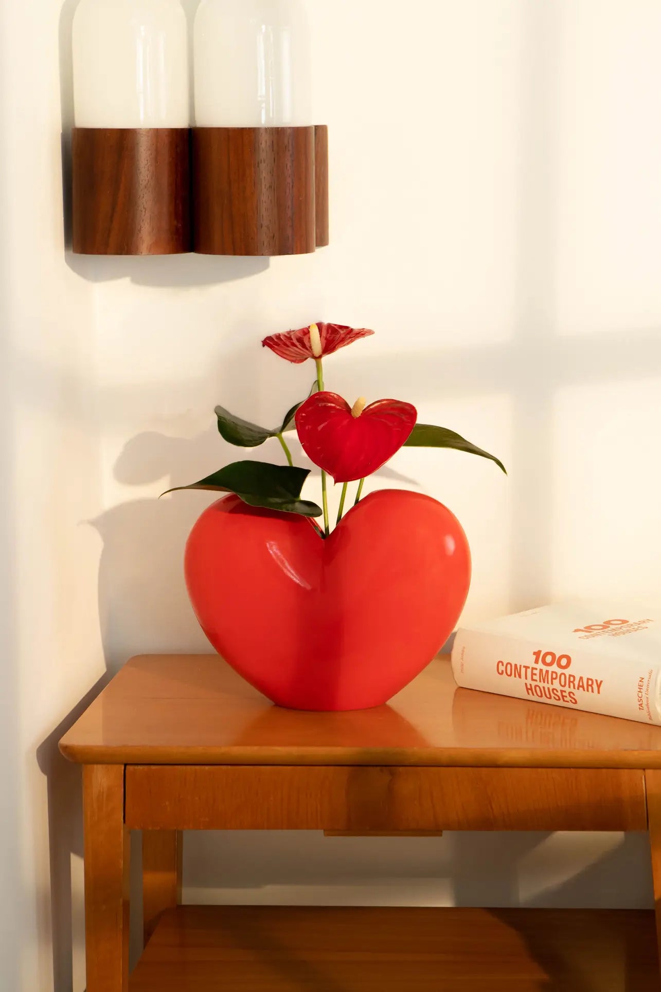 The DOIY Love Vase by DOIY, featuring a red heart shape with red flowers and green leaves, rests on a wooden table next to the book 100 Contemporary Houses, highlighted by warm natural light.