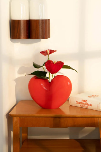 The DOIY Love Vase by DOIY, featuring a red heart shape with red flowers and green leaves, rests on a wooden table next to the book 100 Contemporary Houses, highlighted by warm natural light.