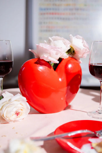 The DOIY Love Vase by DOIY, shaped like a heart, displays white roses on a pink tablecloth beside two glasses of red wine, with a single rose and cutlery nearby. A blurred multicolored chart adds to this display’s sentimental and timeless beauty.