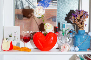 A colorful shelf display features the DOIY Love Vase—a red, heart-shaped vase by DOIY with white roses—next to a wine glass with red liquid, cheese, a blue vase of dried flowers, candles, a postcard, and decor against an art print backdrop.