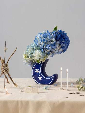 The DOIY Moon Vase by DOIY, featuring a blue ceramic finish and white face design, holds blue hydrangeas on a beige table. Two lit candles, twigs, and petals add to the celestial-inspired scene against a plain gray background.