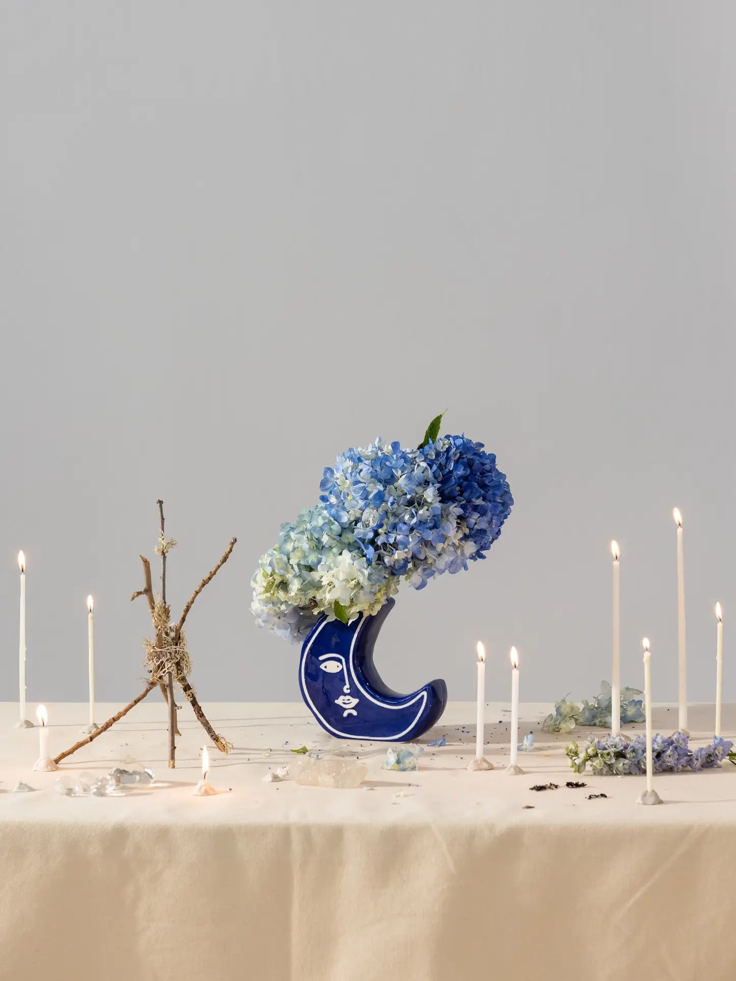 The DOIY Moon Vase by DOIY, filled with blue and white hydrangeas, is displayed on a table with lit white candles, a small branch sculpture, and scattered flower petals against a plain light gray background.