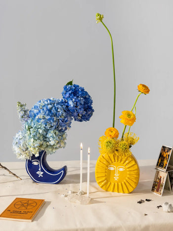 Two celestial-inspired vases by DOIY—a blue Moon Vase with hydrangeas and a yellow sun-themed vase with bright blooms—sit on a table decorated with tarot cards, an orange book, and two lit candles.