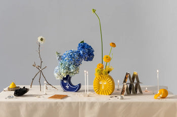 A cream-colored table displays the DOIY Moon Vase by DOIY, adorned with blue and yellow flowers, candles, a branch, a house of cards, crystal pieces, stones, a lemon, and a tarot card—all set against a plain background.