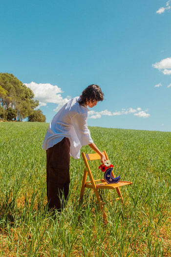 A person in a white shirt and brown pants stands in a green field, placing red flowers in the DOIY Moon Vase by DOIY, which rests on a wooden chair beneath a bright blue sky with scattered clouds.