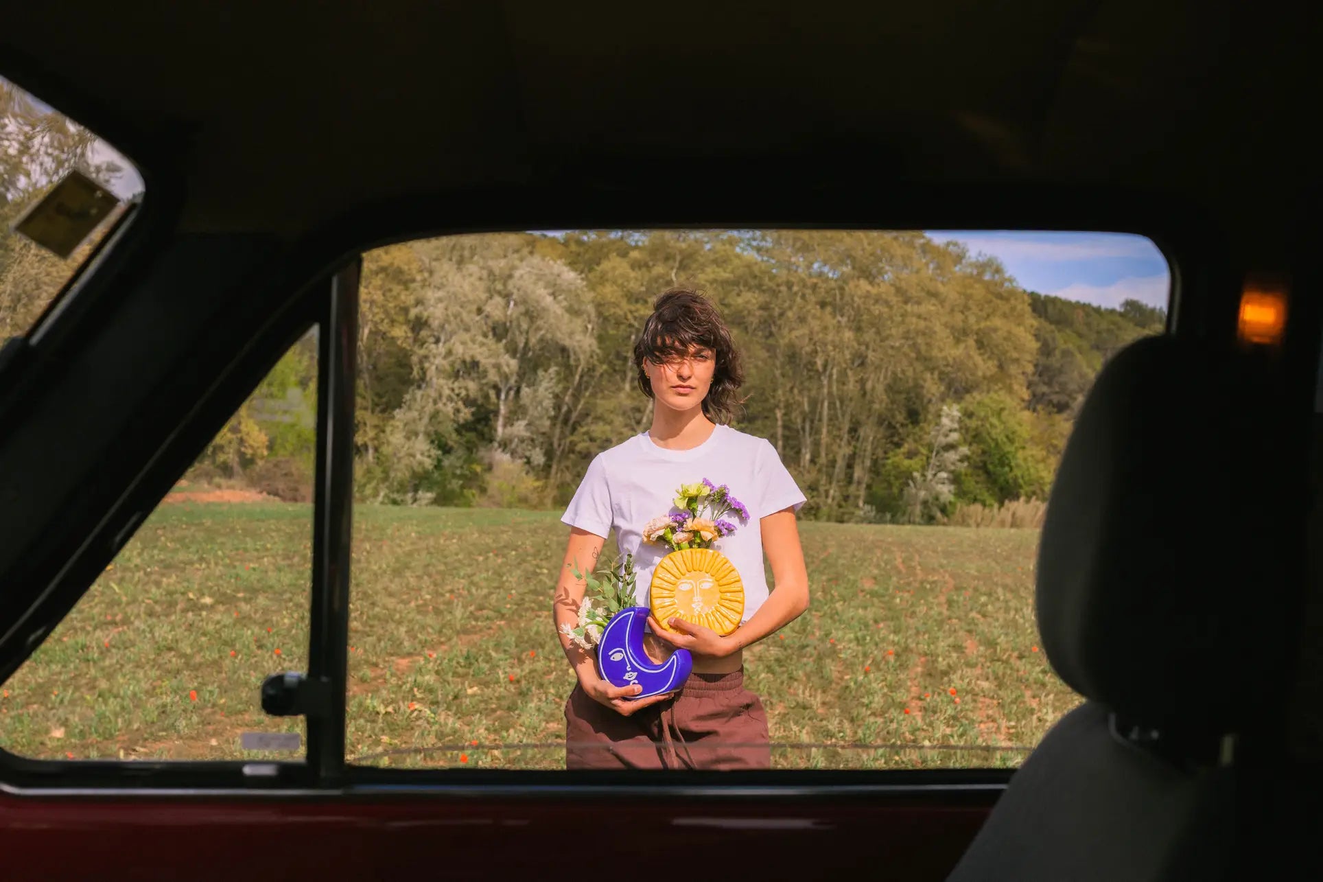 A person with short brown hair and glasses holds the DOIY Moon Vase by DOIY in a field, viewed through a car window, creating a celestial-inspired scene with trees and greenery under a clear sky.
