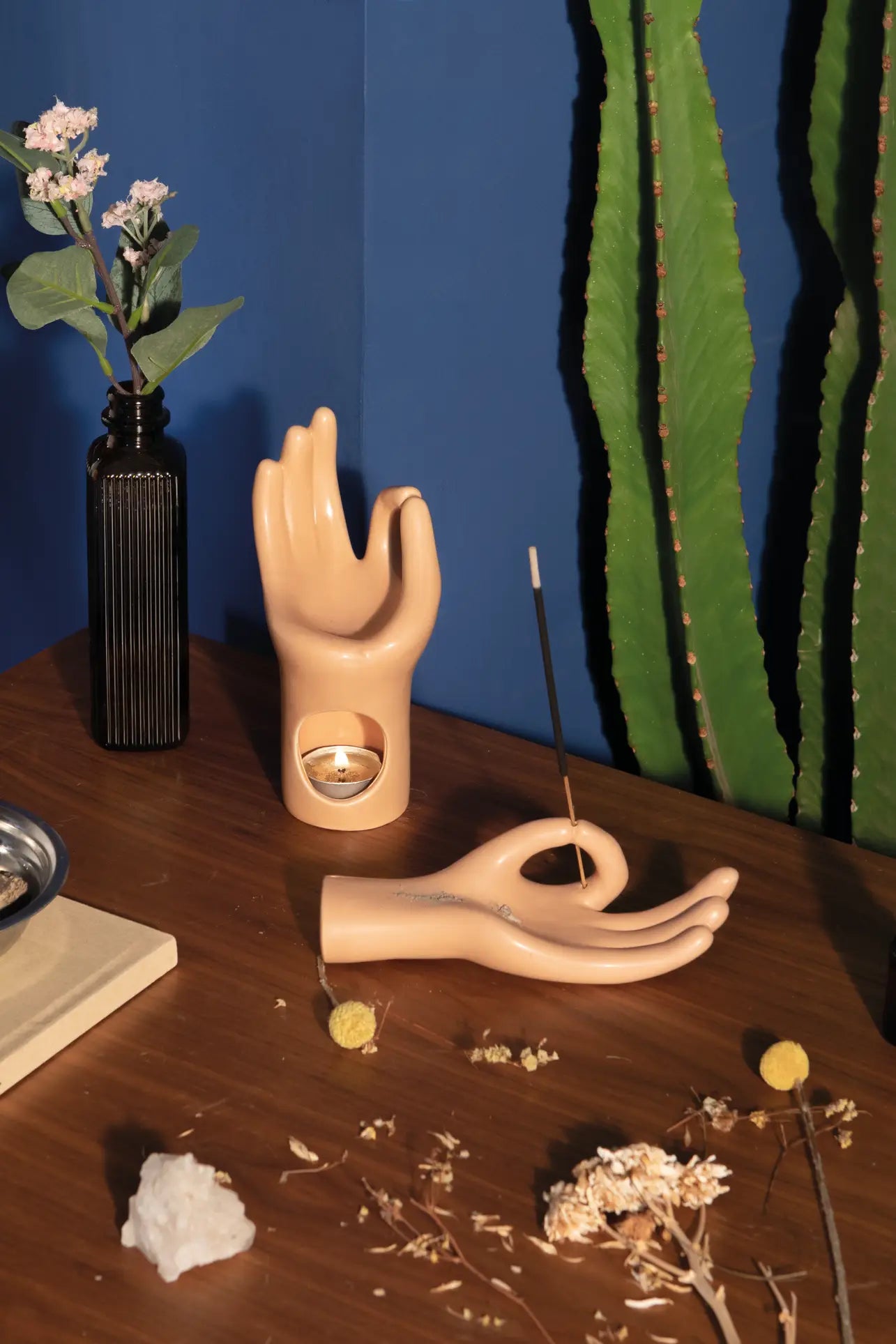 The DOIY Om Meditation Hand Incense Holder by DOIY rests on a wooden table with a tealight, dried flowers, a black vase, crystal, and cactus—ideal mindfulness accents set against a blue wall.