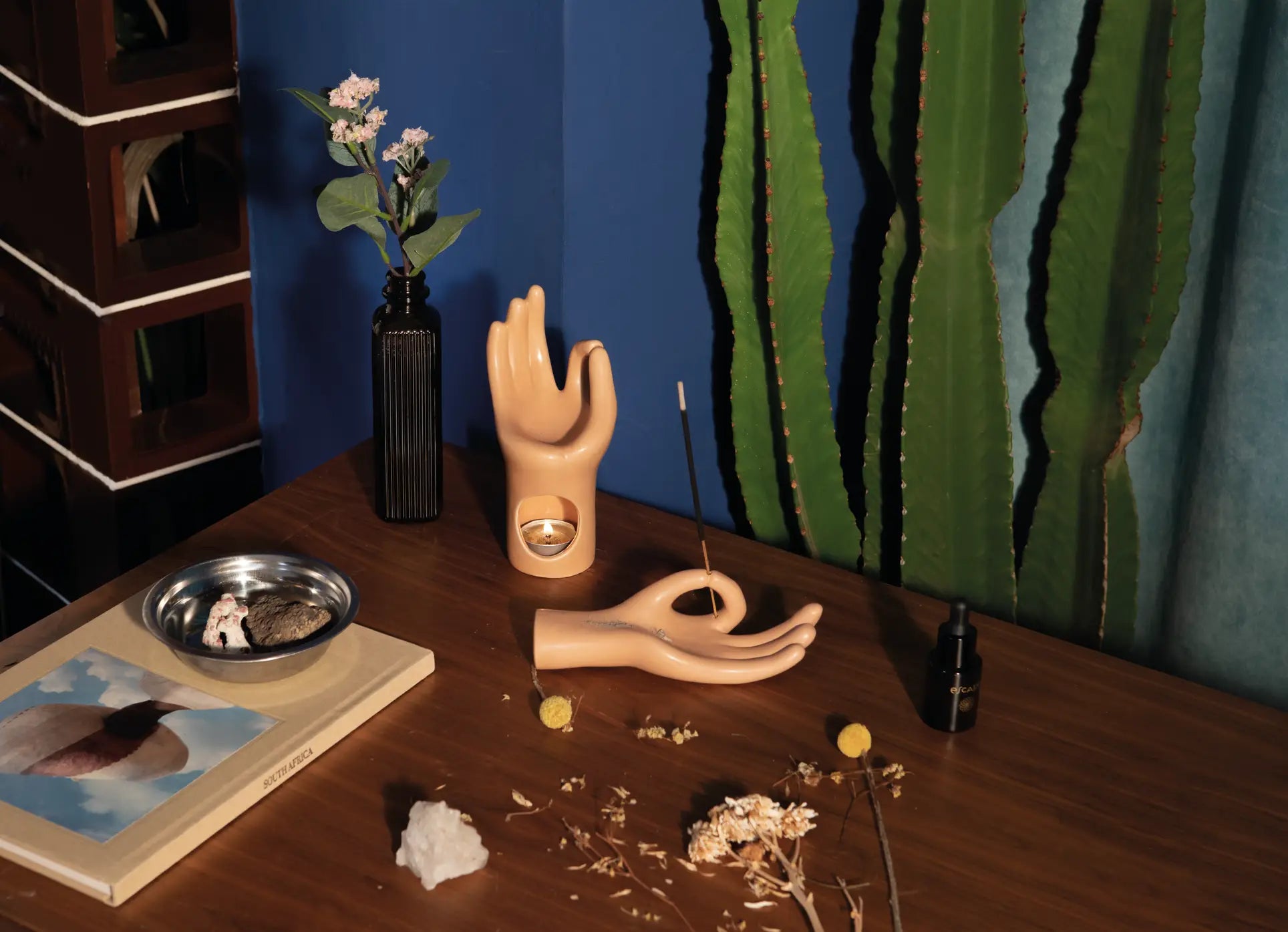 Against a blue wall with green curtains, a wooden table displays the DOIY Om Meditation Hand Incense Holder by DOIY, an incense stick, small flower vases, a crystal, a book, stones on a tray, and a tall cactus for a mindful vibe.