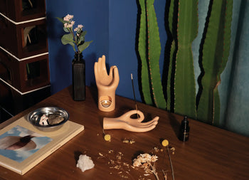 Against a blue wall with green curtains, a wooden table displays the DOIY Om Meditation Hand Incense Holder by DOIY, an incense stick, small flower vases, a crystal, a book, stones on a tray, and a tall cactus for a mindful vibe.