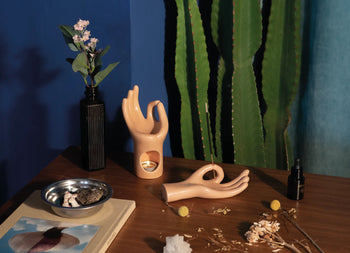 A still life scene with a vase of flowers, two ceramic hand sculptures, the DOIY Om Meditation Hand Incense Holder by DOIY, a metal bowl, a book, dried flowers, a dropper bottle, and a tall cactus against a blue wall on a wooden table.