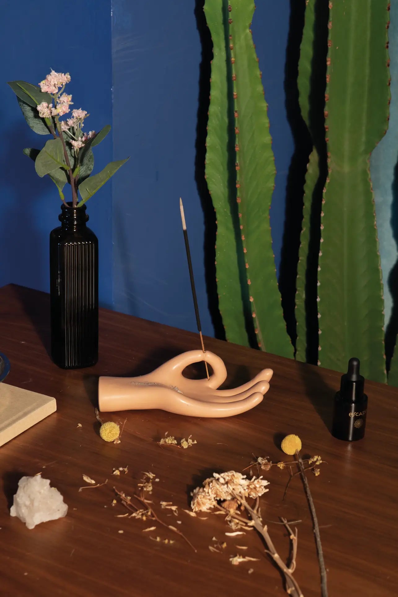 The DOIY Om Meditation Hand Incense Holder by DOIY, a ceramic hand symbolizing mindfulness, holds a burning stick on a wooden table surrounded by dried flowers, greenery in a black vase, a black bottle, crystal, and cactus against a blue wall.