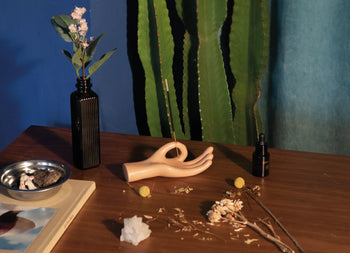 A tabletop featuring the DOIY Om Meditation Hand Incense Holder, a black vase of flowers, dropper bottle, crystal, dried flowers, metal dish, and a book. A tall green cactus stands against a blue backdrop.