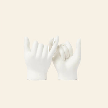 The DOIY Pinky Swear Salt and Pepper Set by DOIY features two white ceramic hand sculptures with interlocked pinky fingers, creating an artistic centerpiece against a light beige background.