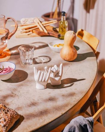 On a round table, food items and a glass of water surround the DOIY Pinky Swear Salt and Pepper Set—a ceramic sculpture by DOIY resembling two hands making pinky promises—bathed in sunlight that casts striking shadows.