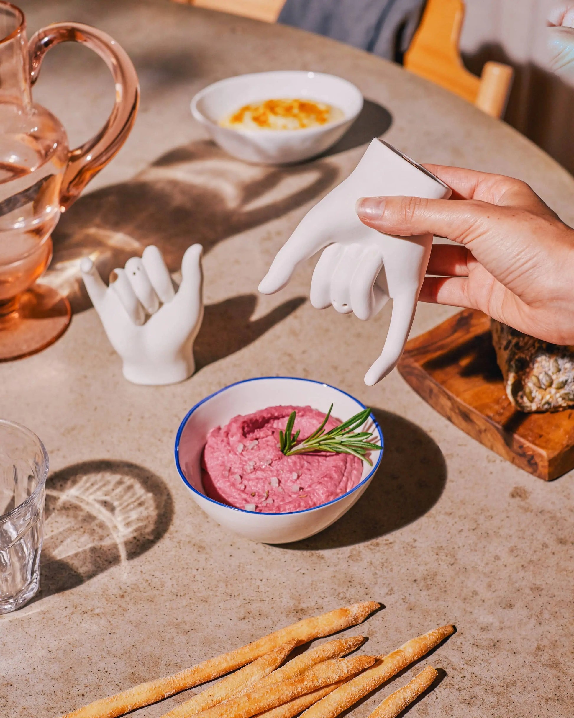 A hand holds a DOIY Pinky Swear Salt and Pepper Set—a white ceramic hand-shaped piece—above a bowl of pink dip with rosemary, surrounded by breadsticks, glassware, and another DOIY ceramic hand on the table.