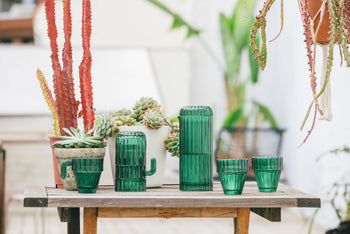 A wooden table displays the DOIY Saguaro Glasses set of 6—stackable cactus-shaped glasses by DOIY—alongside two potted saguaro cacti and other succulents, with hanging and potted plants brightening the outdoor decor.