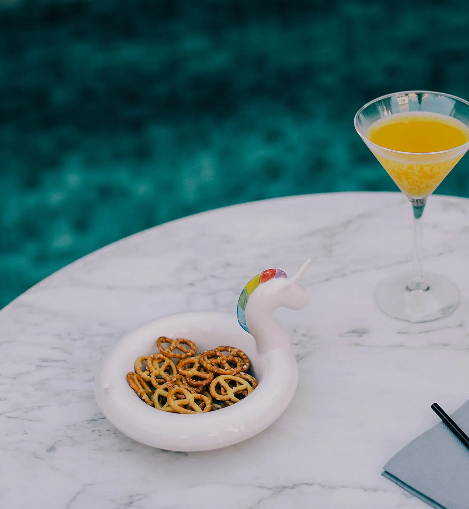 By the pool, a marble table holds a DOIY Unicorn Floatie Bowl by DOIY filled with pretzels, a martini glass with an orange-yellow drink, plus a folded gray napkin and black straw.