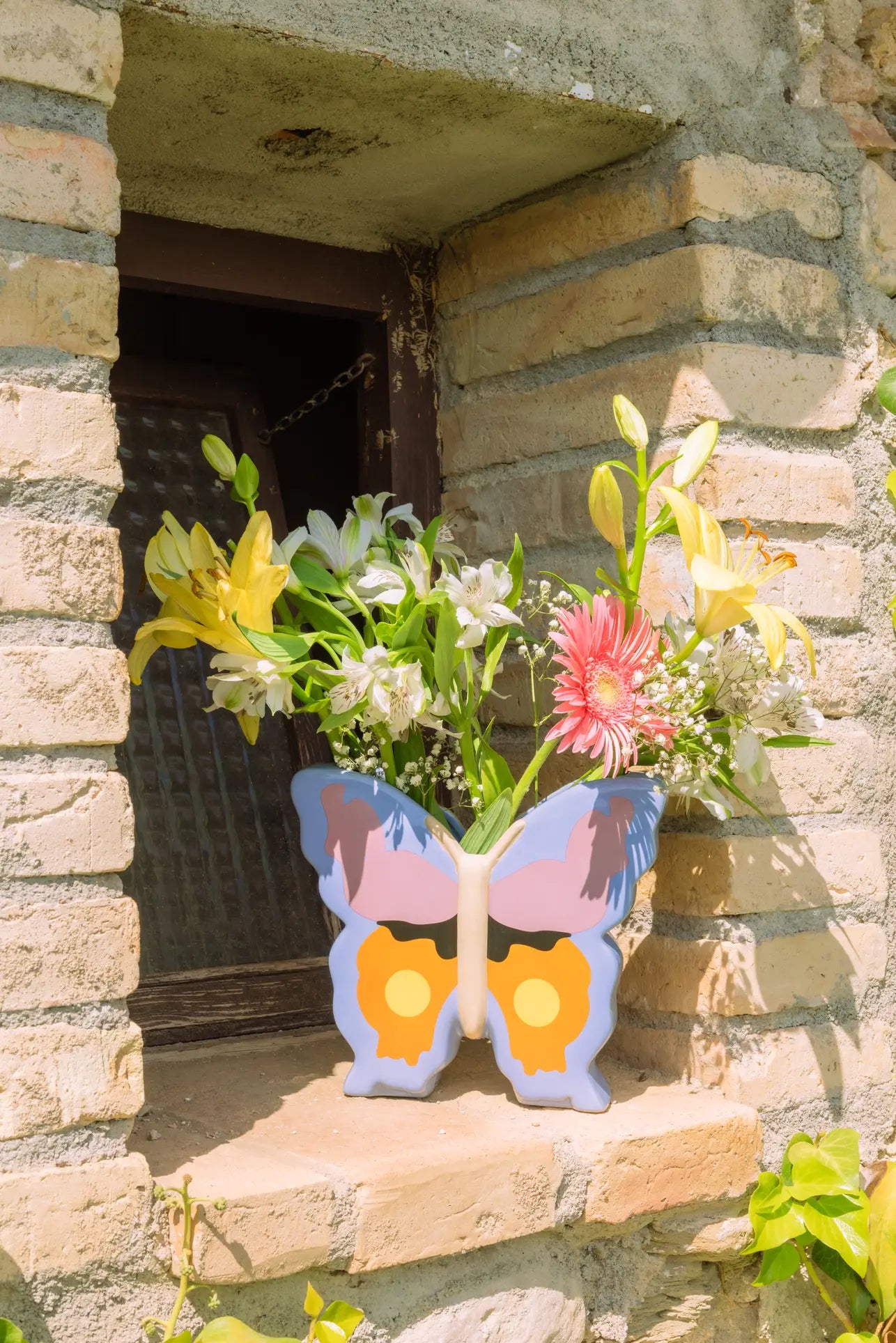 The DOIY Woodland Butterfly Vase by DOIY, filled with yellow, white, and pink flowers, rests on a stone windowsill outside a rustic brick building, adding charming cottage style to the scene.