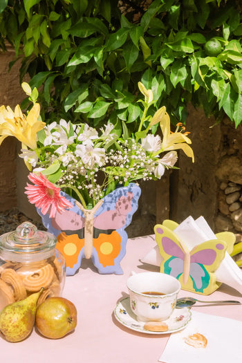 An outdoor table features a DOIY Woodland Butterfly Vase filled with flowers, a cup of coffee, pears, and cookies in a jar. Nearby, a butterfly silhouette napkin holder complements the look and adds to the cottage ambiance.
