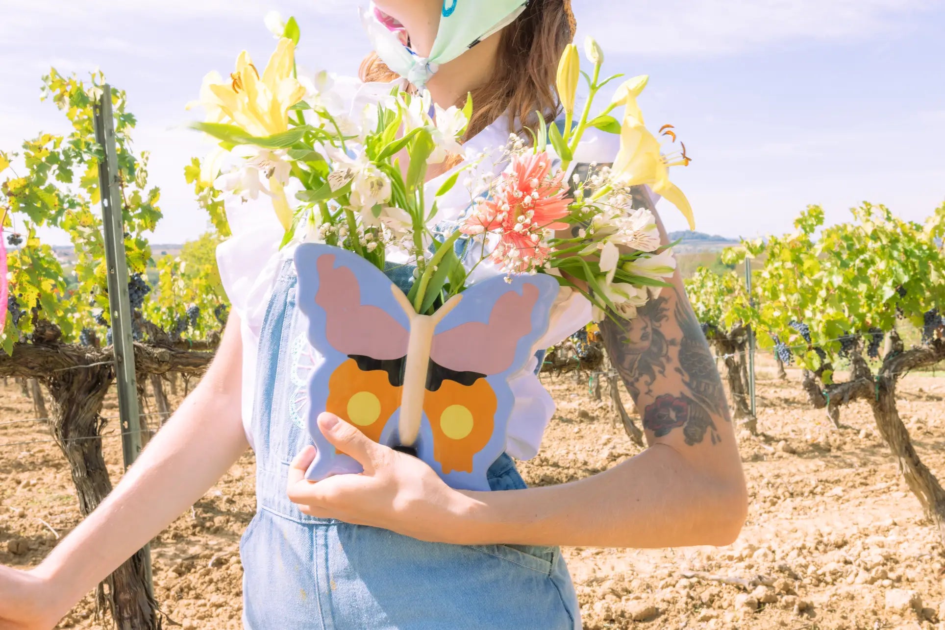A person in denim overalls holds the DOIY Woodland Butterfly Vase by DOIY, filled with colorful flowers. They stand in a sunny vineyard, their tattooed arm and face mask visible, creating a gentle cottage-inspired scene.