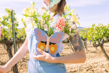 A person in denim overalls holds the DOIY Woodland Butterfly Vase by DOIY, filled with colorful flowers. They stand in a sunny vineyard, their tattooed arm and face mask visible, creating a gentle cottage-inspired scene.