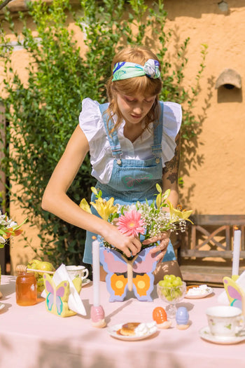 A woman in a headband and overalls arranges flowers in the DOIY Woodland Butterfly Vase by DOIY on a pink outdoor table with candles, fruit, drinks, and teacups, evoking a charming cottage vibe amid greenery and a peach-colored wall.
