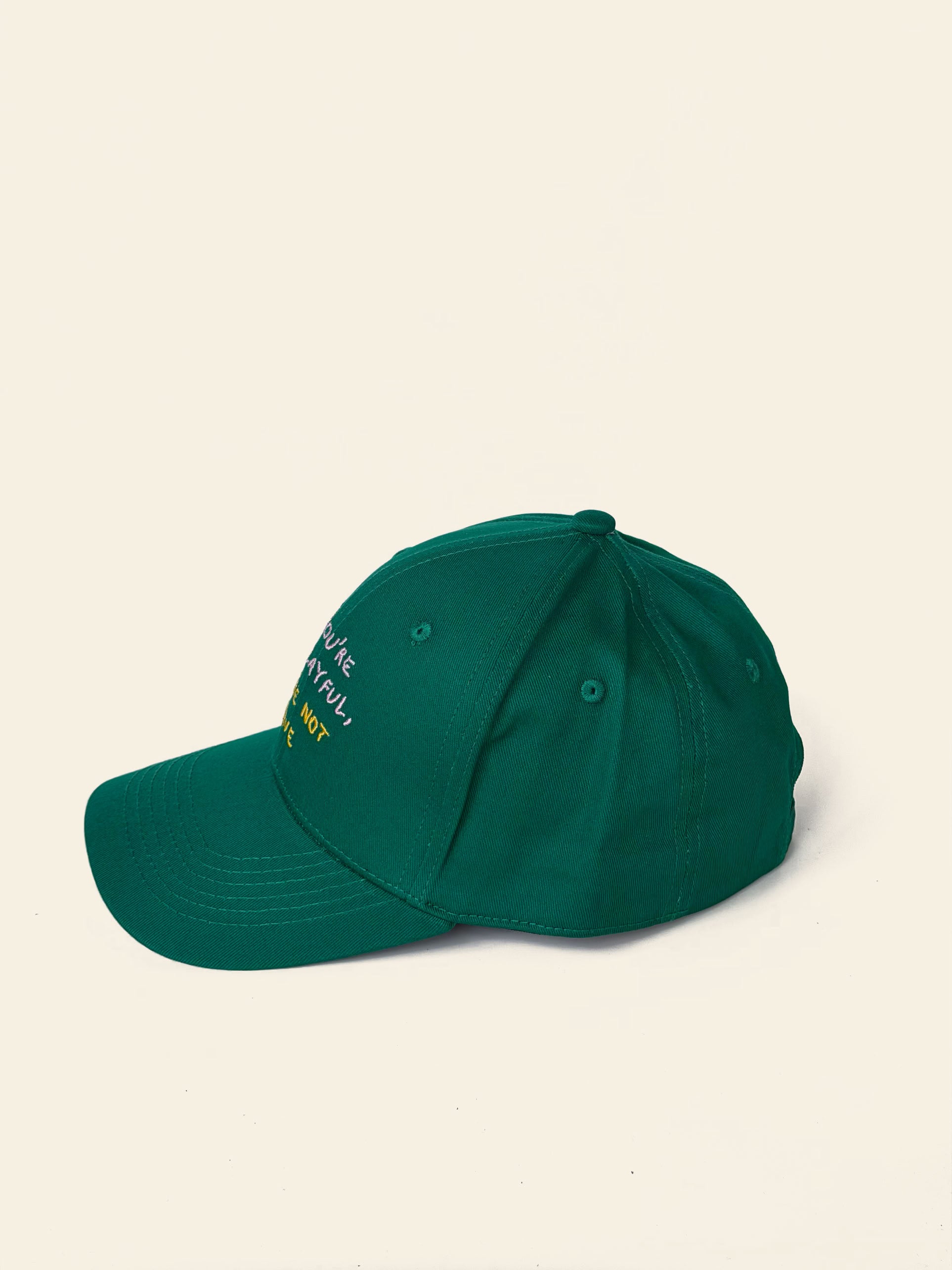 The David Hockney If You’re Not Playful Cap—a dark green cotton cap by David Hockney, featuring white and yellow embroidered text on the side—is showcased against a plain off-white background and captures the artist’s famously spirited, playful style.
