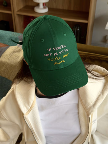A person wearing the David Hockney If You’re Not Playful Cap by David Hockney—a green cotton cap with yellow and white embroidered text that reads, “IF YOU’RE NOT PLAYFUL, YOU’RE NOT ALIVE”—is sitting indoors on a couch, their face partially obscured by the playful brim, reminiscent of David Hockney’s iconic style.
