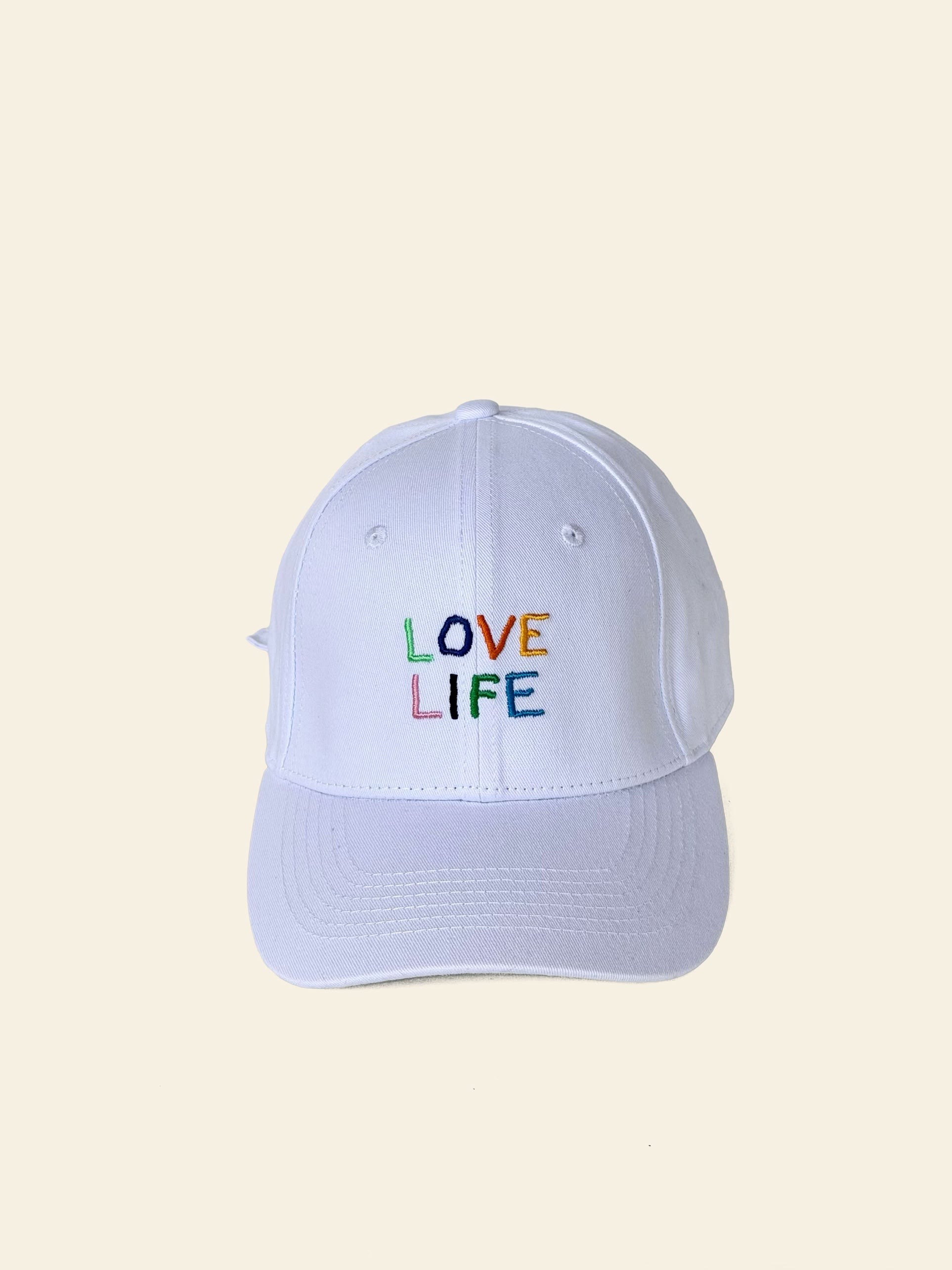 A David Hockney Love Life Cap by David Hockney, featuring the words “LOVE LIFE” embroidered in colorful, rainbow letters on the front of a white cotton cap, set against a plain light background.