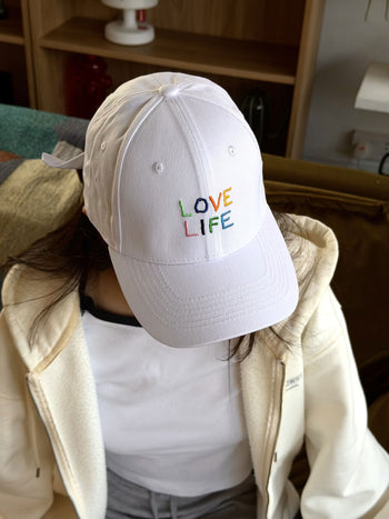 A person wearing the David Hockney Love Life Cap—a white cotton hat with LOVE LIFE embroidered in colorful letters by David Hockney—sits indoors, dressed in a white jacket over a black-and-white shirt.
