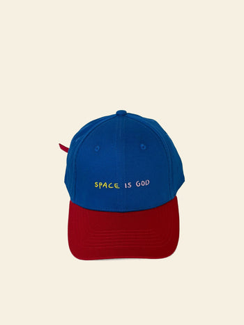 The David Hockney Space Is God Cap by David Hockney is a blue two-tone cotton cap with a red brim, featuring “SPACE IS GOD” embroidered in yellow and white on the front.